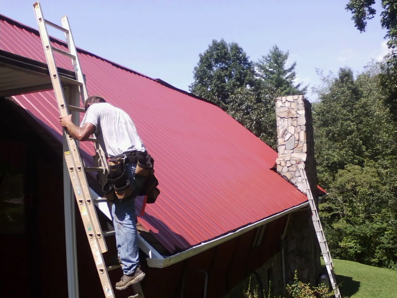 Roofer on a ladder beside a red metal roof and stone chimney — Soffit & Fascia Repair by Roofing Brothers in Scott Bar